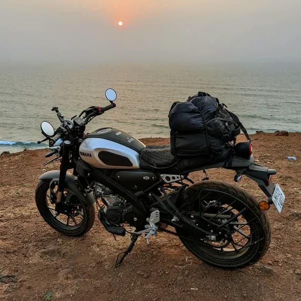 A Yamaha XSR motorcycle, loaded with a large black bag, is parked on a reddish-brown dirt path overlooking the ocean. A small, orange sun is visible low in the hazy sky above the water, creating a soft, warm glow.