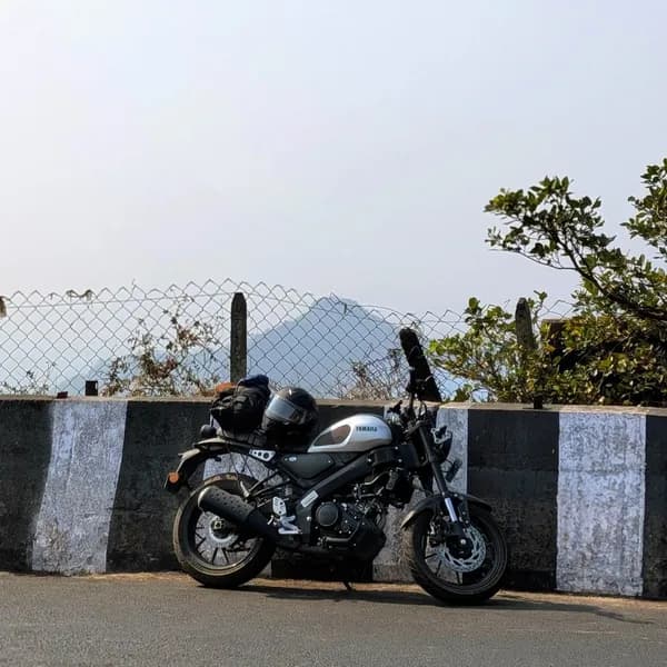 A silver-tanked Yamaha XSR motorcycle, equipped with a helmet and rear luggage, is parked on an asphalt road next to a black and white striped concrete barrier. Beyond a chain-link fence, hazy mountains rise under a bright sky, with green foliage on the right.