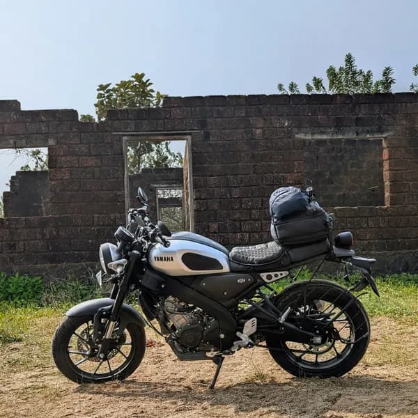 A silver and black Yamaha XSR motorcycle, loaded with a rear luggage bag, is parked on sandy ground before a weathered brick wall with empty window frames, all beneath a clear blue sky.